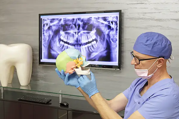 A dentist wearing scrubs and gloves holding a TMJ model in front of an X-ray screen, explaining jaw anatomy and dysfunction.
